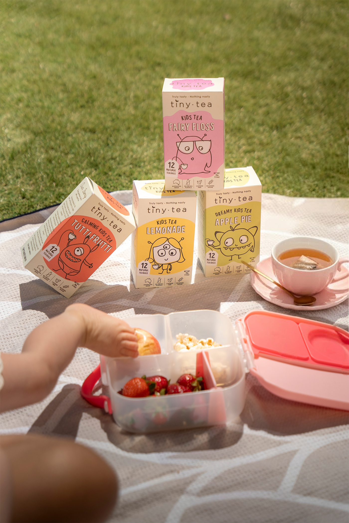 Tiny Tea boxes with a child's hand reaching for snacks on a picnic blanket outdoors.